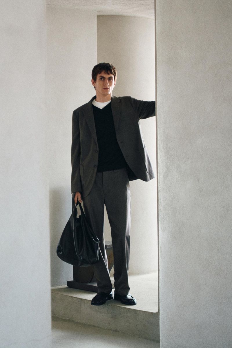 Man wearing a grey suit jacket and trousers holding a black leather bag while standing in an architectural interior