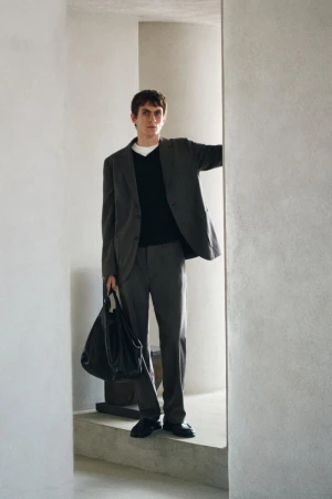 Man wearing a grey suit jacket and trousers holding a black leather bag while standing in an architectural interior
