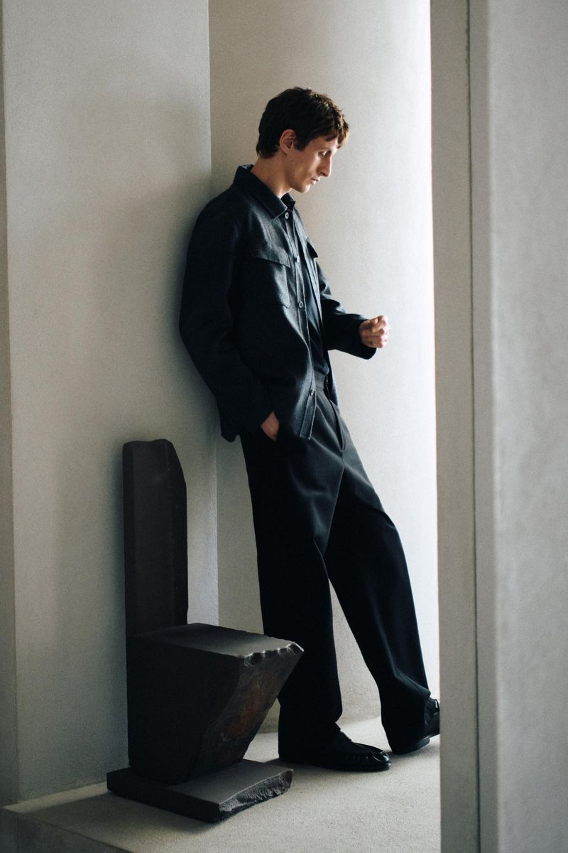 Man wearing a dark jacket and trousers leaning against a wall next to a sculptural chair