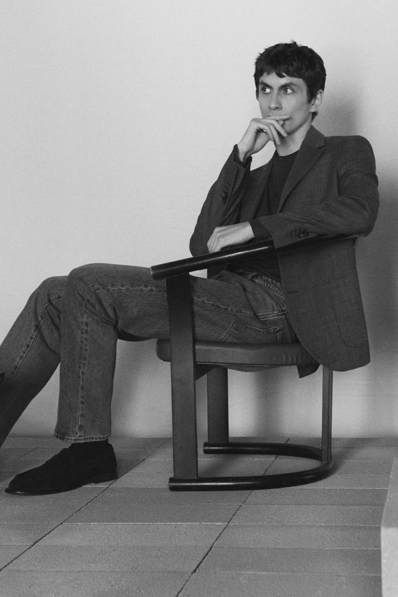Man seated on a black chair wearing a blazer and jeans against a plain studio background.