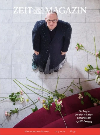High-angle portrait of David Sedaris wearing a dark coat standing on a light marble floor near scattered flowers and a burgundy edge