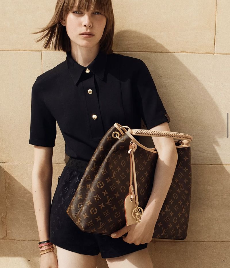 Fashion shot of a woman holding a brown Louis Vuitton handbag wearing a black top and shorts against a light brown wall.