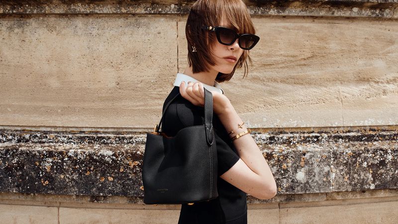Thea Almqvist holding a black leather bucket bag while wearing dark sunglasses and a black collared top against a stone wall.