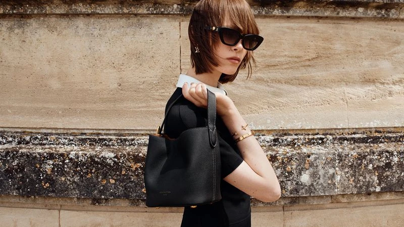 Thea Almqvist holding a black leather bucket bag while wearing dark sunglasses and a black collared top against a stone wall.