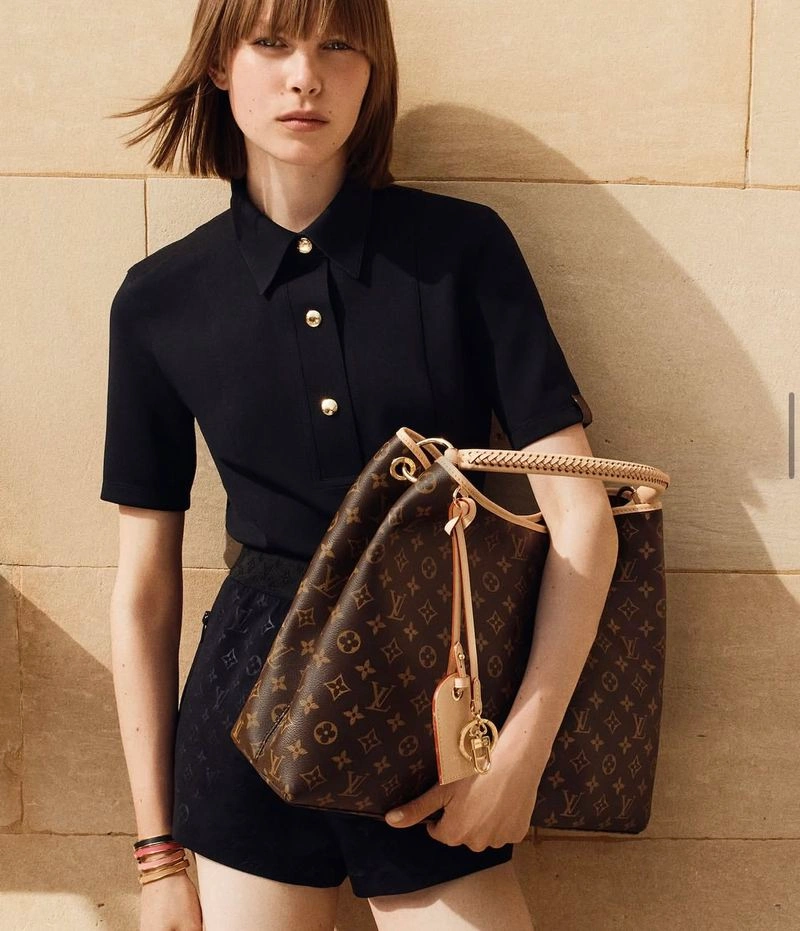 Model with bob haircut wearing black collared shirt holding a monogram tote bag against a stone wall