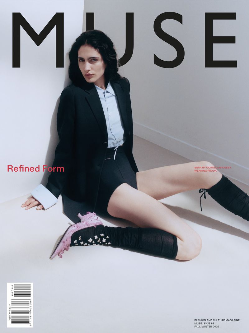 Model Sara seated on a white floor against a corner wall wearing a black blazer, light blue shirt, black shorts, and pink heels.