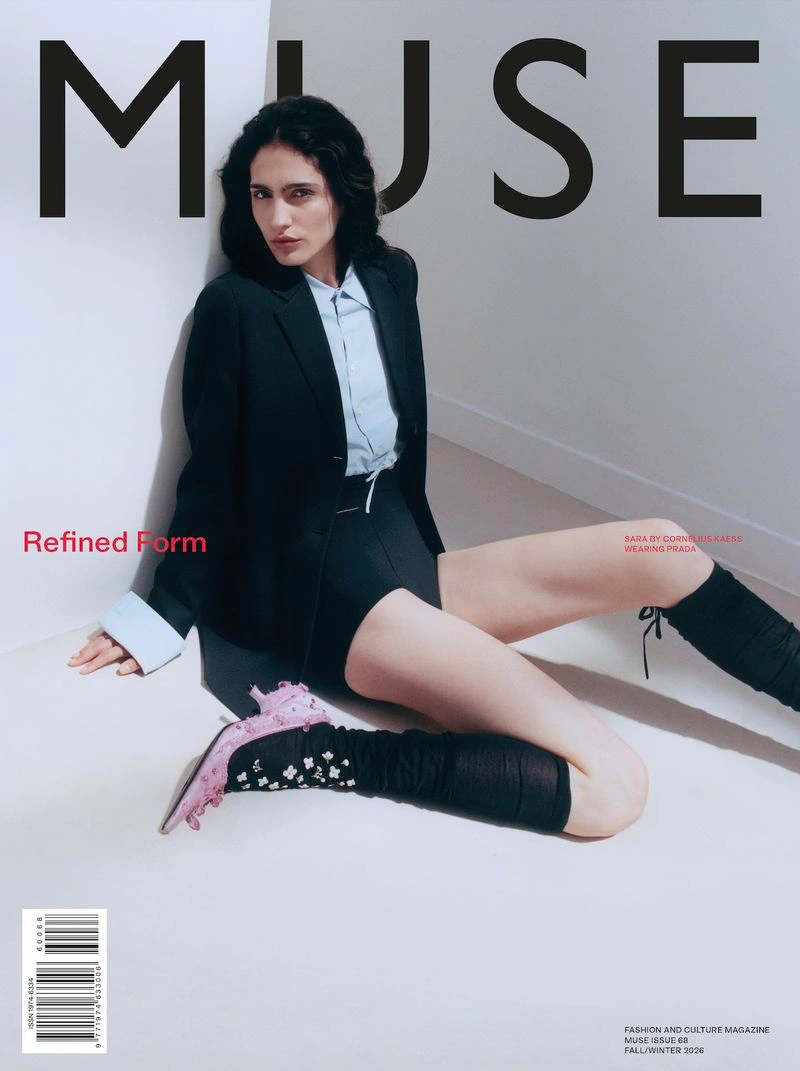 Model Sara seated on a white floor against a corner wall wearing a black blazer, light blue shirt, black shorts, and pink heels.