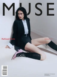 Model Sara seated on a white floor against a corner wall wearing a black blazer, light blue shirt, black shorts, and pink heels.