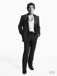 Tom Cruise standing against a plain white background wearing a dark tailored suit jacket, trousers, and a white button-down shirt.