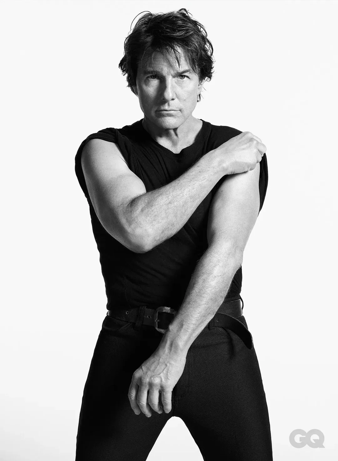 Tom Cruise in a black t-shirt and dark trousers posing against a plain white background