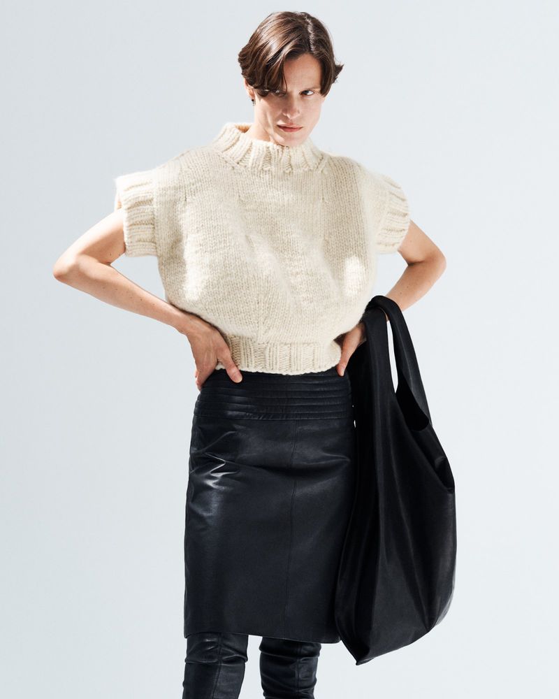 Model wearing a textured cream knit top and black leather skirt holding a large black handbag against a white background