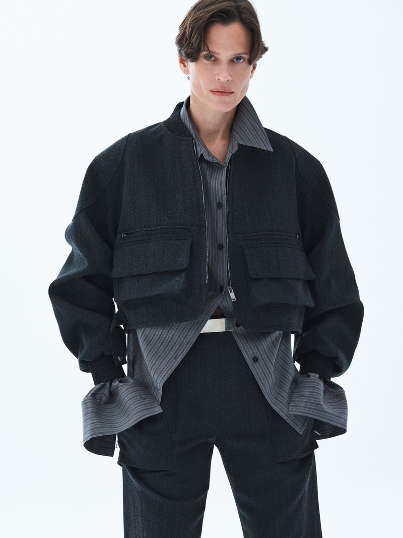 Model wearing a dark utility bomber jacket and striped shirt against a plain white studio background