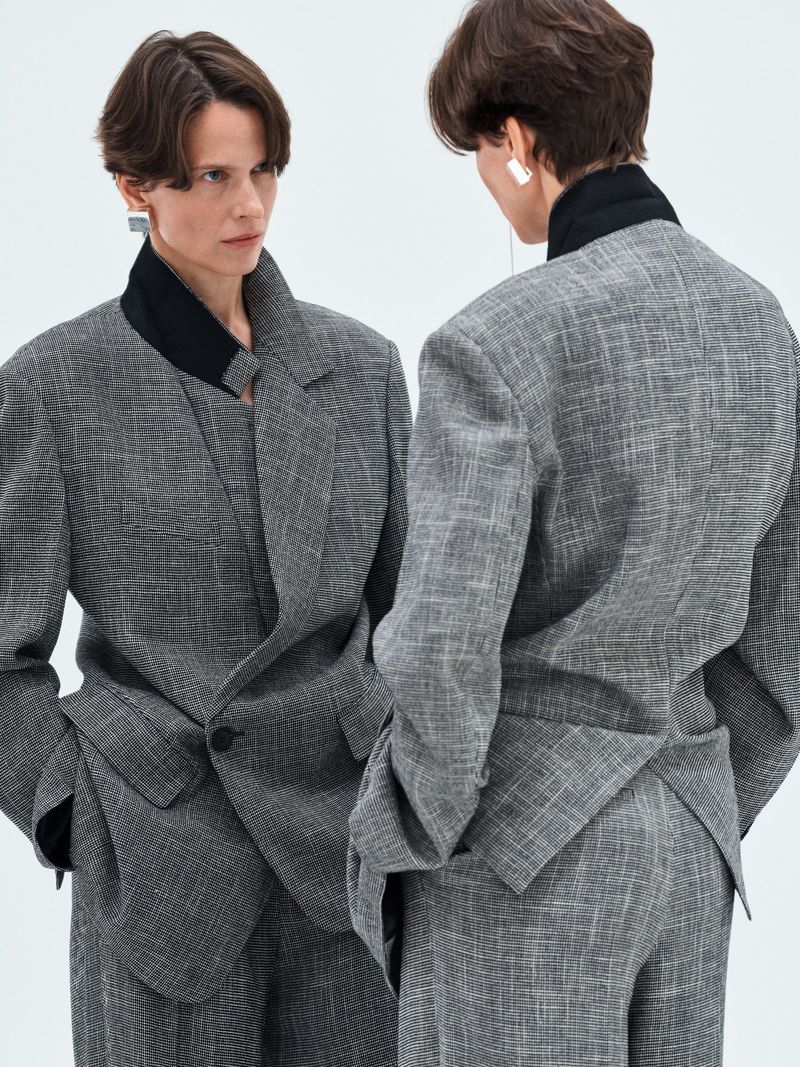 Model wearing a grey textured blazer and matching trousers reflected in a mirror against a plain white background.