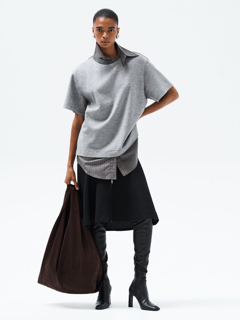 Model wearing a grey short-sleeve top and black skirt with knee-high boots holding a large dark bag against a white backdrop.