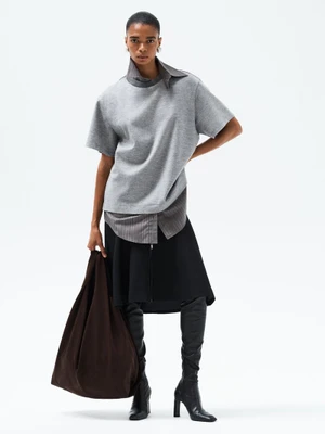 Model wearing a grey short-sleeve top and black skirt with knee-high boots holding a large dark bag against a white backdrop.