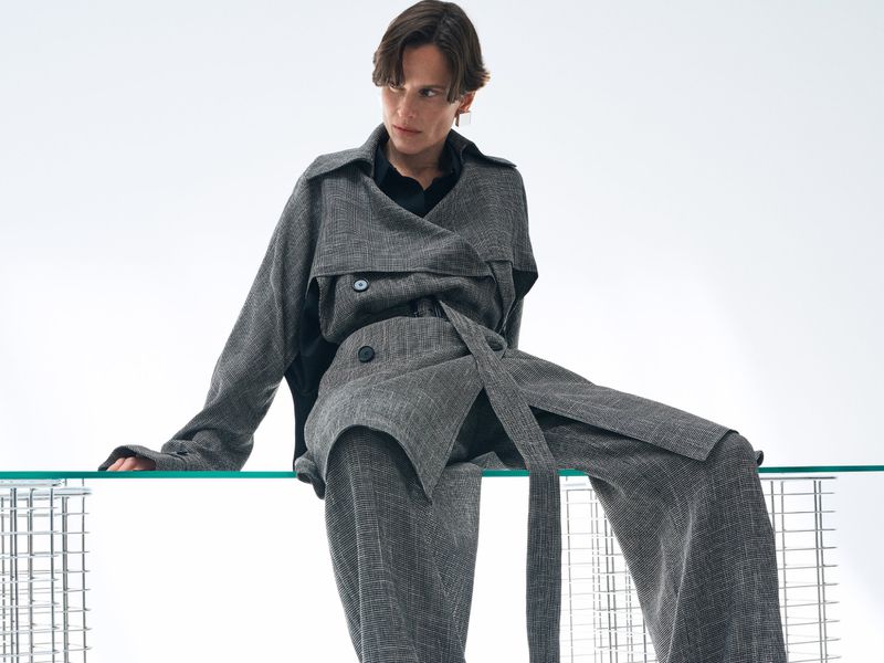 Model seated on a glass edge wearing a grey textured coat and matching trousers against a plain white background
