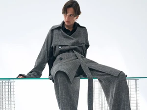 Model seated on a glass edge wearing a grey textured coat and matching trousers against a plain white background