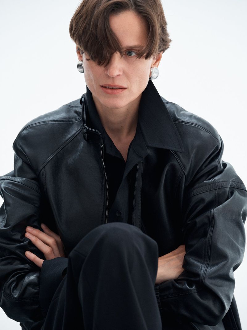 Model in black leather jacket wearing silver earrings against a plain white seamless studio background.