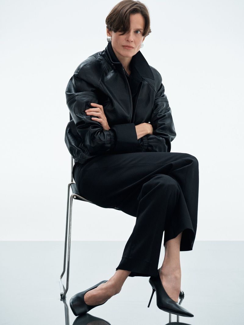 Model wearing a black leather jacket and trousers seated on a metal chair against a white seamless background