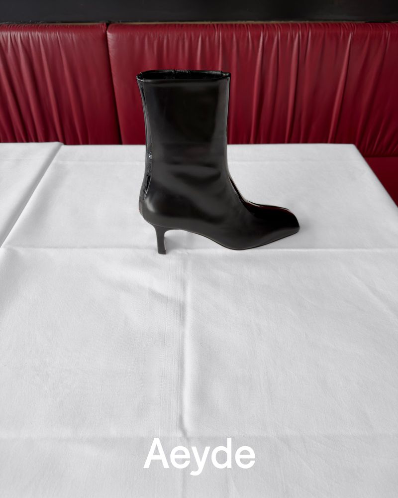 Black leather ankle boot with a kitten heel resting on a white tablecloth against a red banquette for Aeyde Fall 26