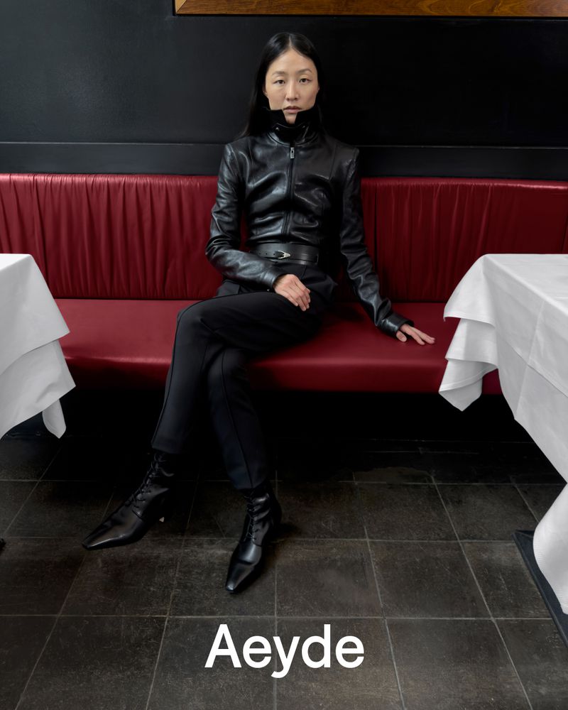 Model seated on a red banquette in a restaurant wearing a black leather jacket and boots for Aeyde Fall 26