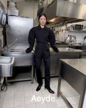 Model wearing a black jacket and trousers standing inside a commercial kitchen with stainless steel counters.