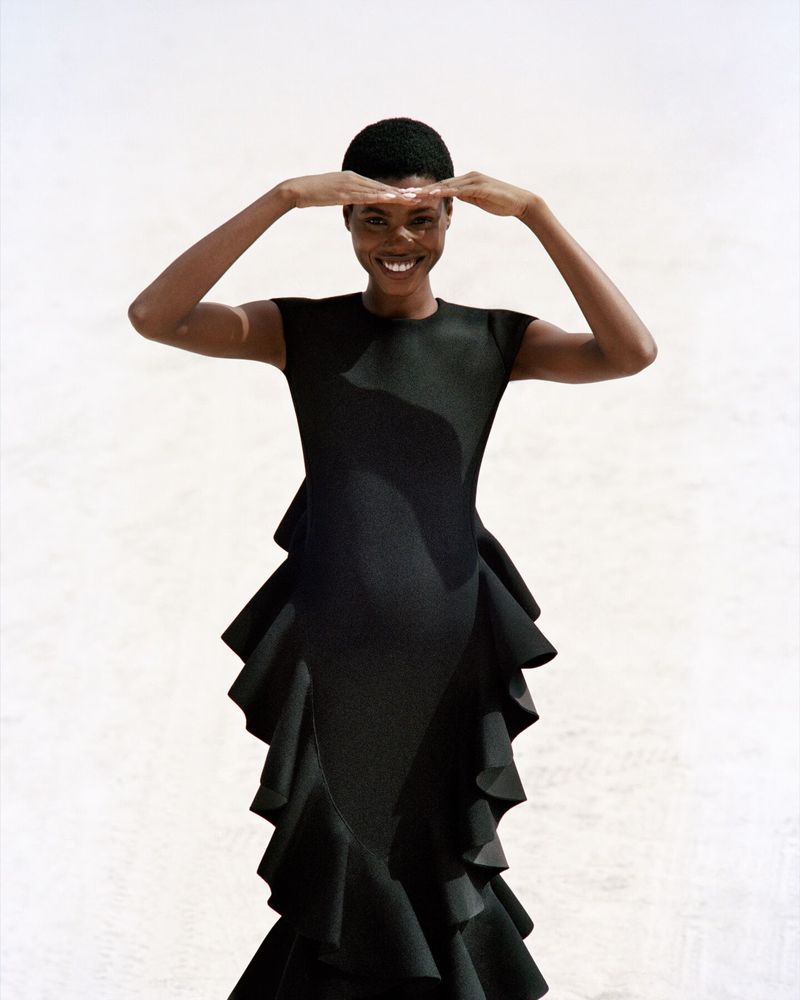 Model in a black dress with ruffled skirt shading eyes against a plain light background