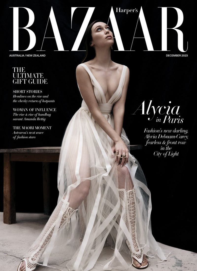 Fashion editorial cover featuring a young woman in a sheer white dress and gladiator-style sandals, gazing upward against a dark background.