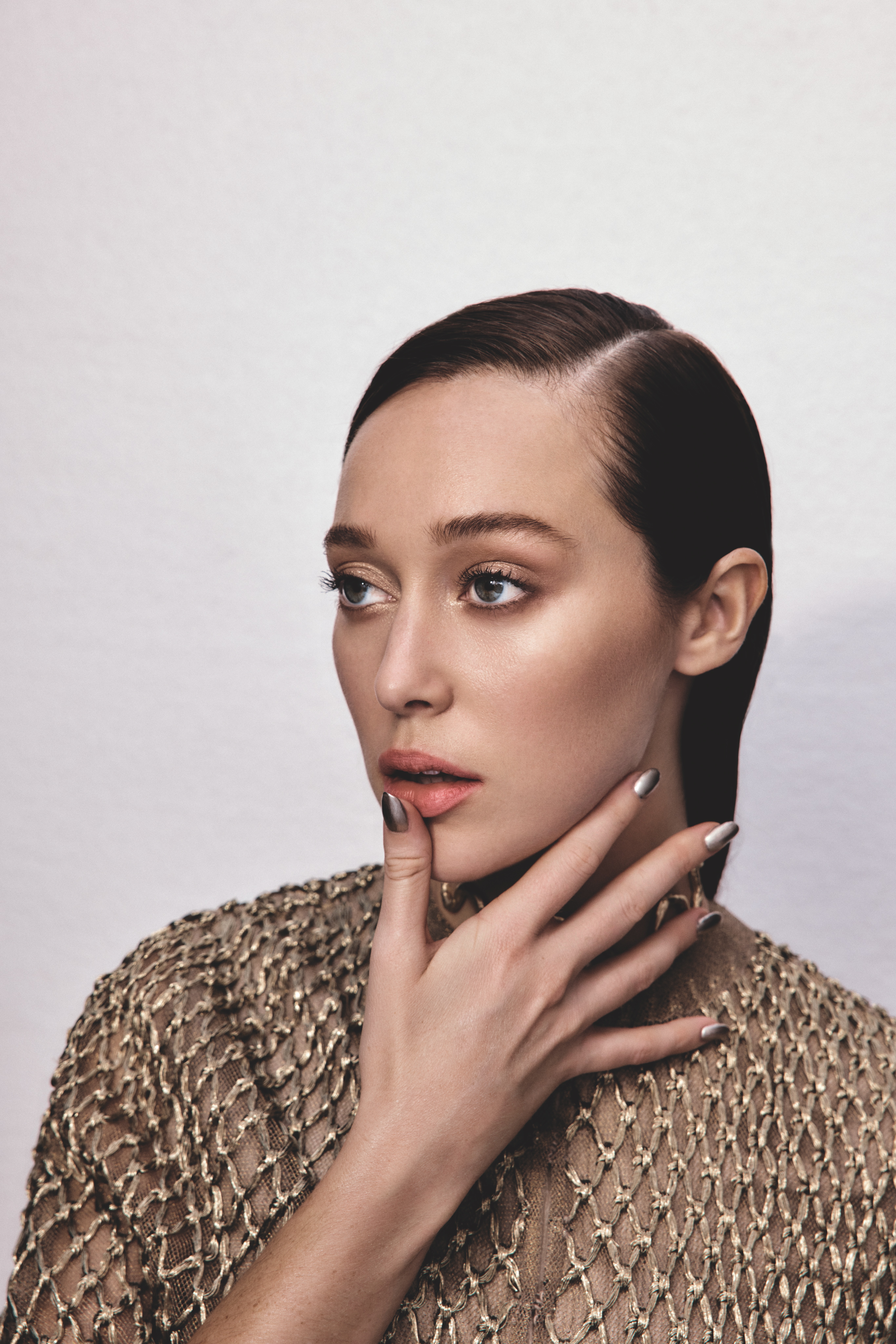 Close-up portrait of a woman with sleek, dark hair, wearing a gold, woven top, gazing off-camera with a hand near her face in a photo studio.
