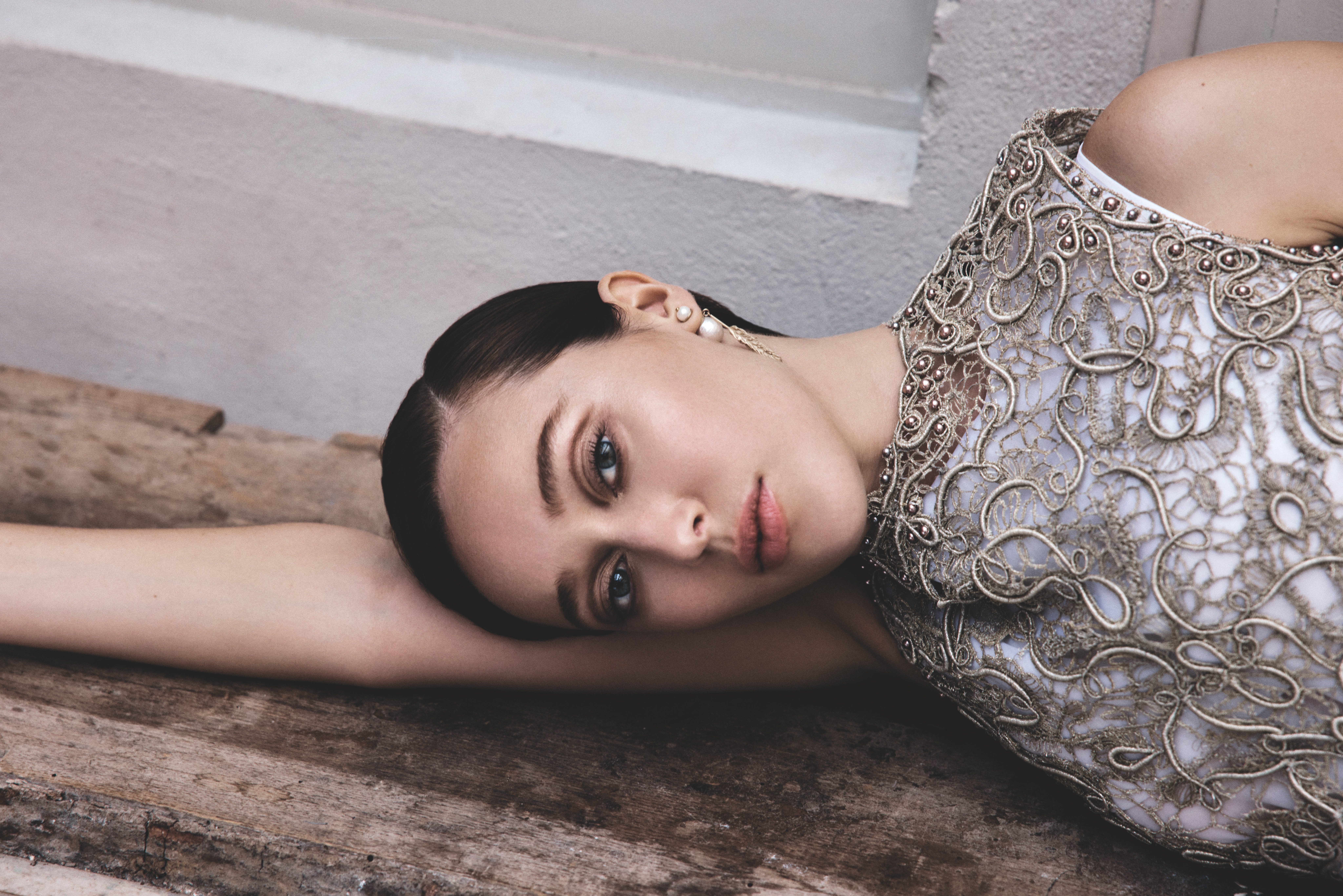 Close-up studio shot of a dark-haired woman in an ornate silver-grey dress, resting on a wooden surface, staring directly at the camera.