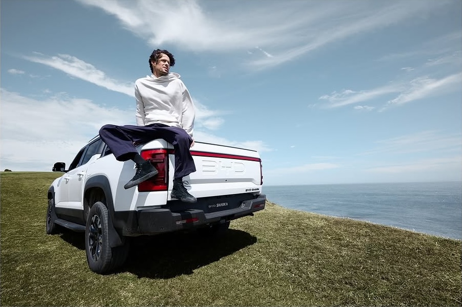 Adult male sits atop a white BYD Shark pickup truck parked on a grassy hill overlooking the ocean, with a blue sky backdrop.