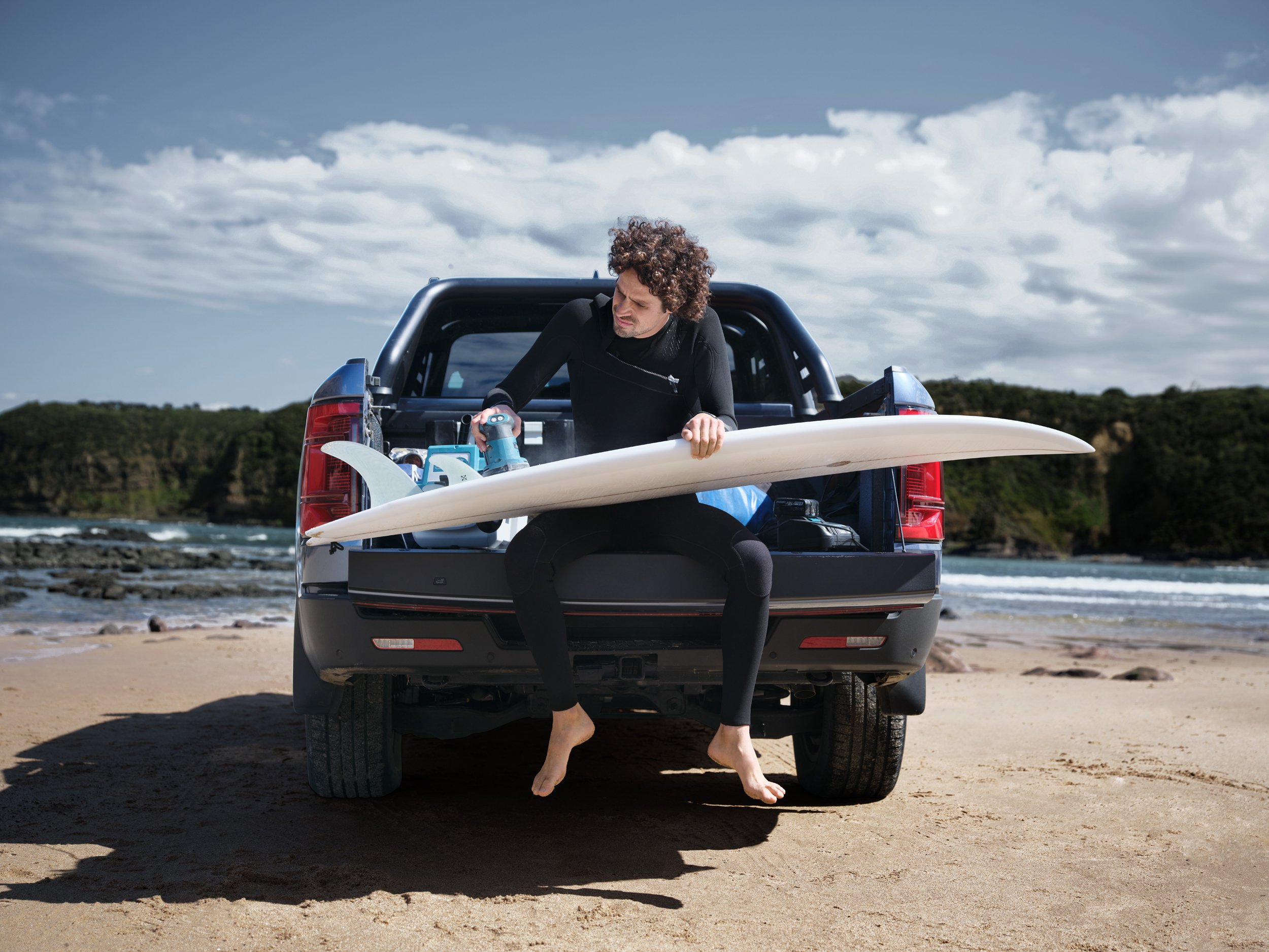 A surfer in a wetsuit sits in the back of a pick-up truck with his surfboard on a beach with a cloudy sky and ocean background.