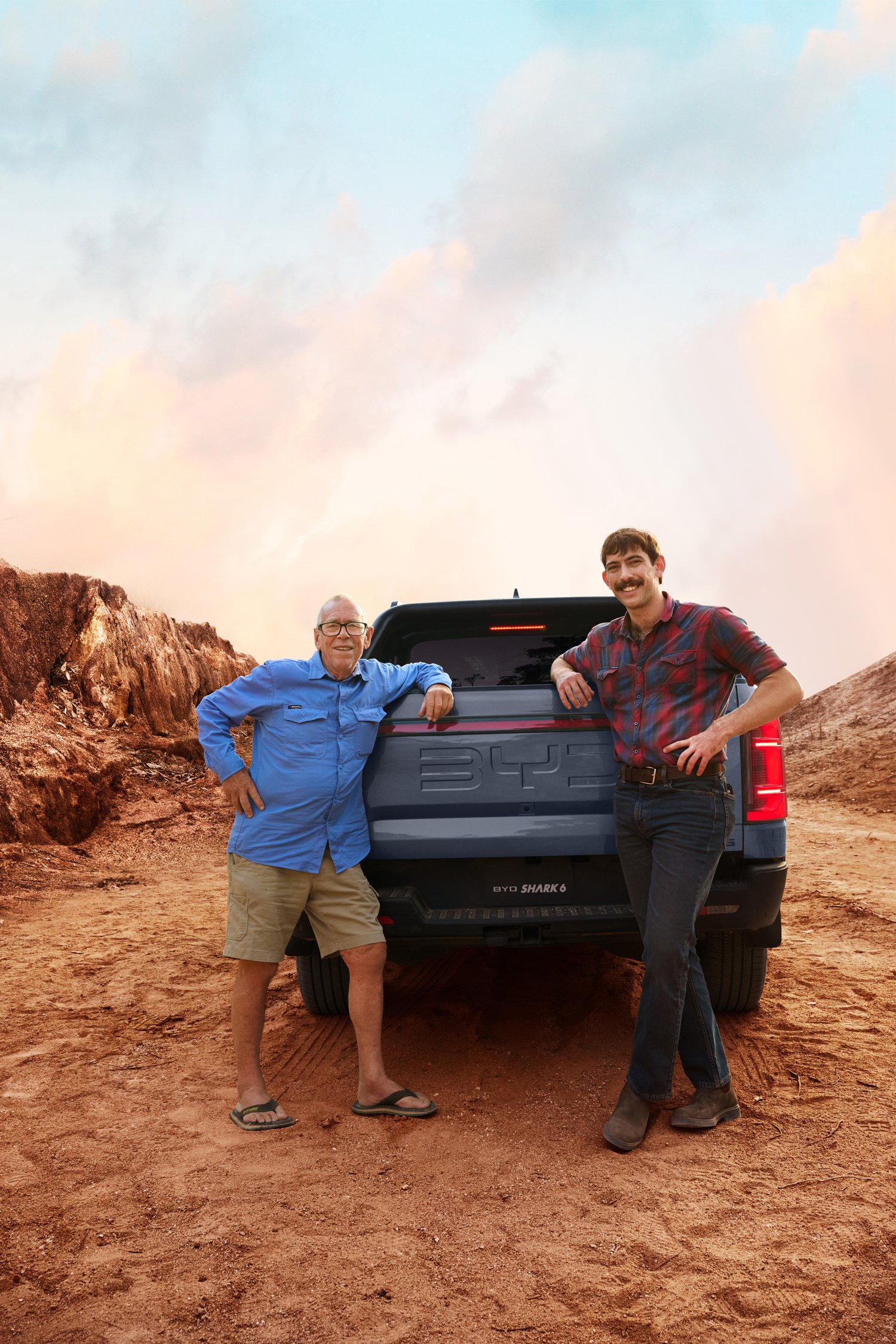 Two men pose by a truck on a dirt road with desert-like landscape and partly cloudy sky at sunset or sunrise, creating a scenic outdoor scene.