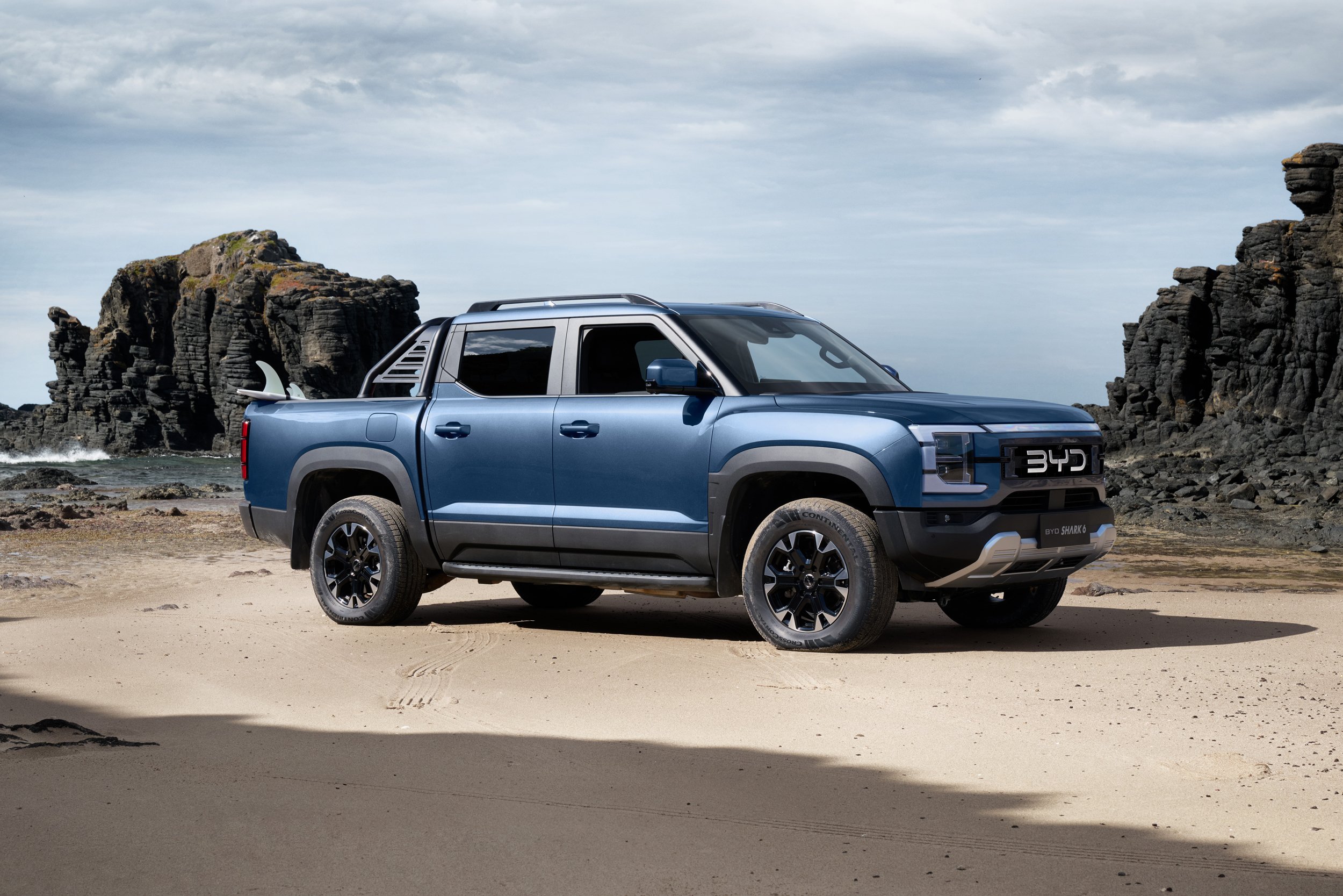 Blue BYD electric pickup truck parked on a sandy beach with a surfboard in the truck bed and rocky cliffs in the background.