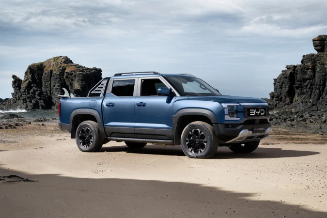 Blue BYD electric pickup truck parked on a sandy beach with a surfboard in the truck bed and rocky cliffs in the background.