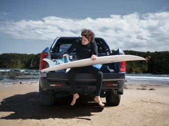 A man in a black wetsuit sitting on the tailgate of a dark pickup truck on a sandy beach holding a surfboard.