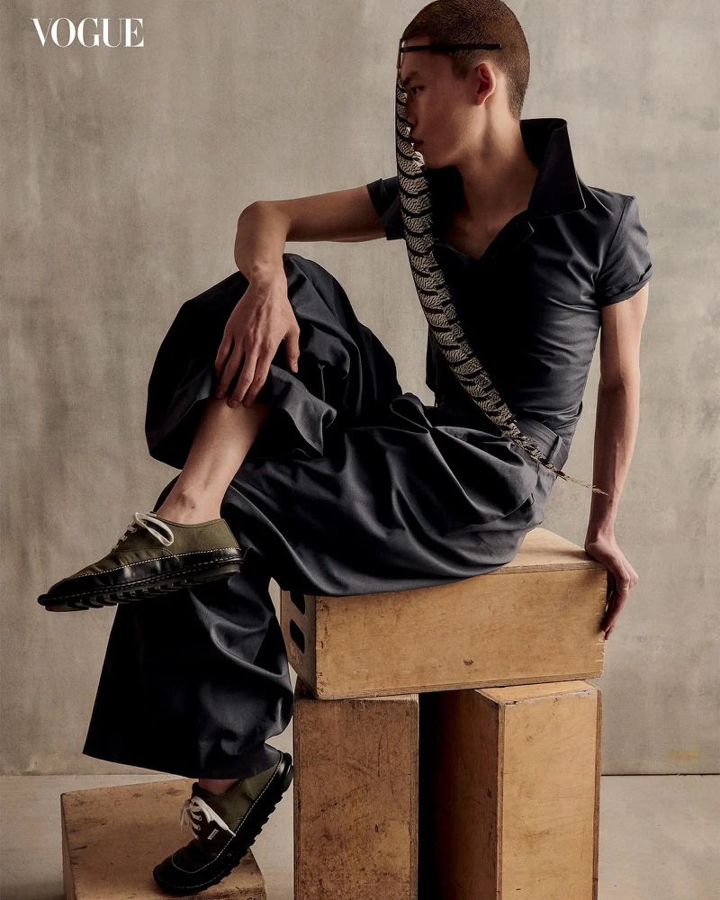 Fashion portrait of a young adult in dark wide-leg pants and matching top sitting on wooden boxes with a feather accessory.