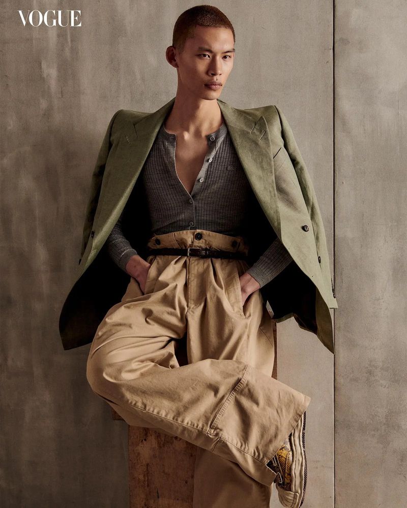 Fashion editorial shot: Asian male model in olive blazer and khaki trousers poses indoors for Vogue against a neutral wall.