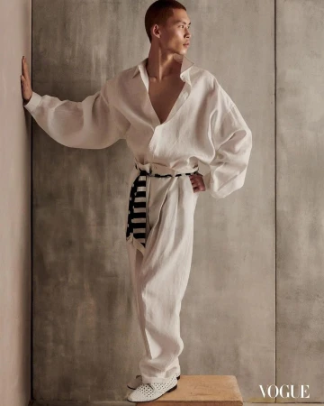 An adult male model wearing a white linen outfit with a striped belt poses in a studio with a painted backdrop and minimal props.