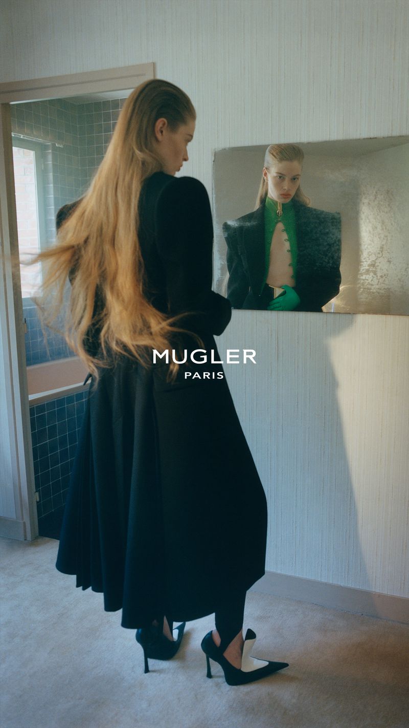 Model in a black coat looking into a mirror in a room, capturing the Mugler fall/winter 26 campaign.