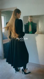 Model in a black coat looking into a mirror in a room, capturing the Mugler fall/winter 26 campaign.