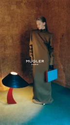 Model in long brown structured dress holding a blue box near a mushroom lamp against a textured cork wall