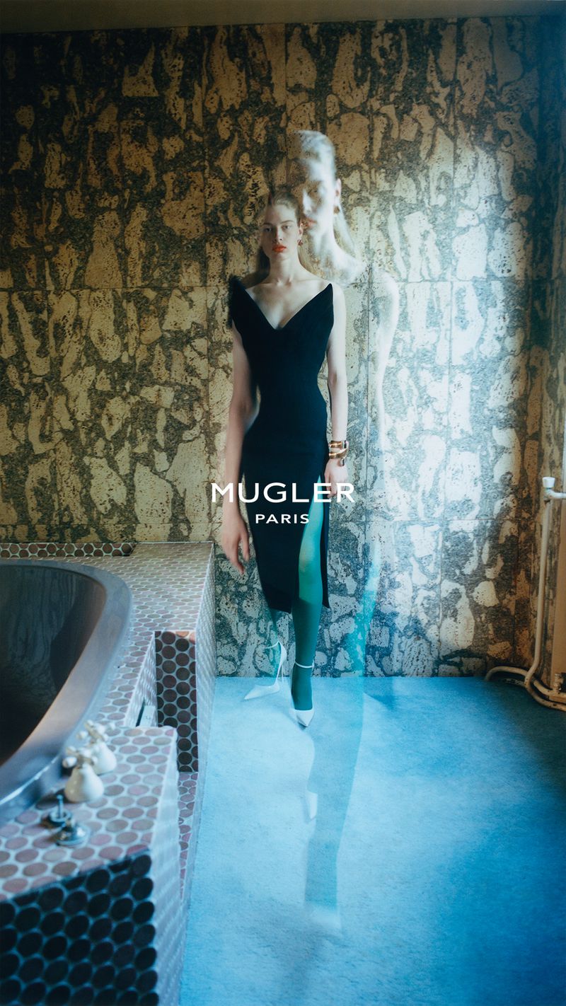 Model in a black dress and green stockings standing indoors with a large textured wall and bathtub.