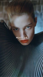Close-up portrait of a model with slicked-back blonde hair, vibrant orange lipstick, and structured grey pleated attire.