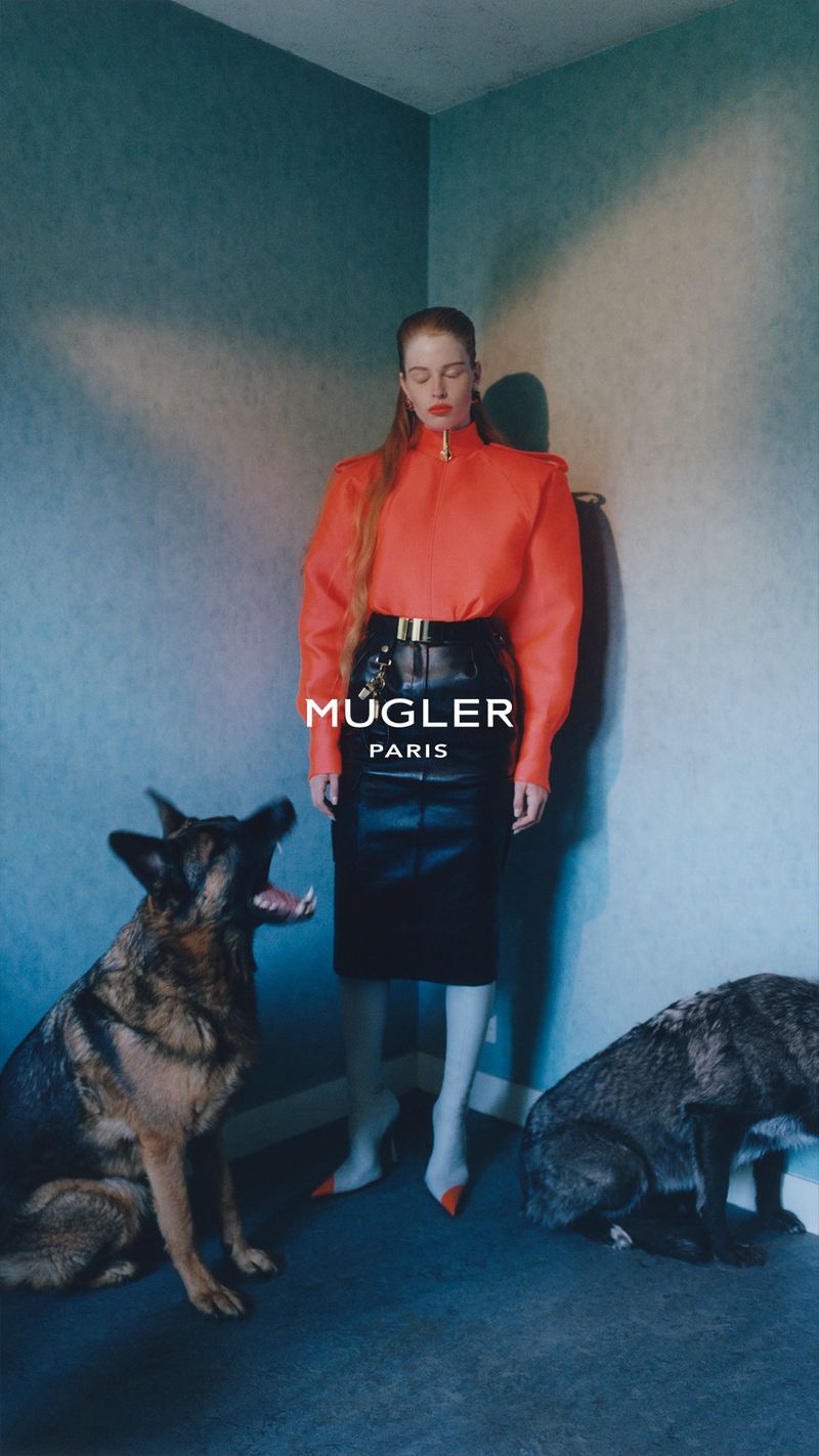 Model in red top and black skirt posing in a room corner with two German Shepherd dogs