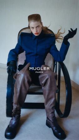 Model seated in a cane-backed chair wearing a blue shirt and tall brown leather boots for Mugler F/W 26