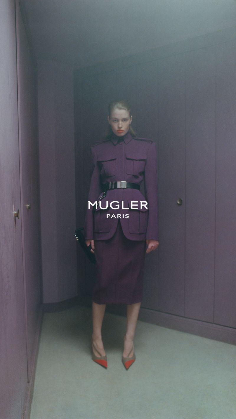 Model standing in a structured plum suit with a belt inside a room with fitted purple wardrobes.