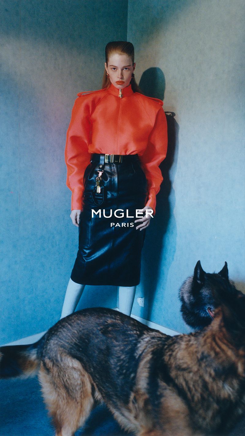 Model in a bright orange structured top and black leather skirt standing in a corner with two dogs in the foreground