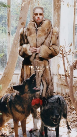 Model wearing a large fur coat and sunglasses with two German Shepherd dogs in a greenhouse setting for a Mugler campaign.