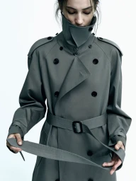 Model in grey belted trench coat holding a belt strap against a bright white background